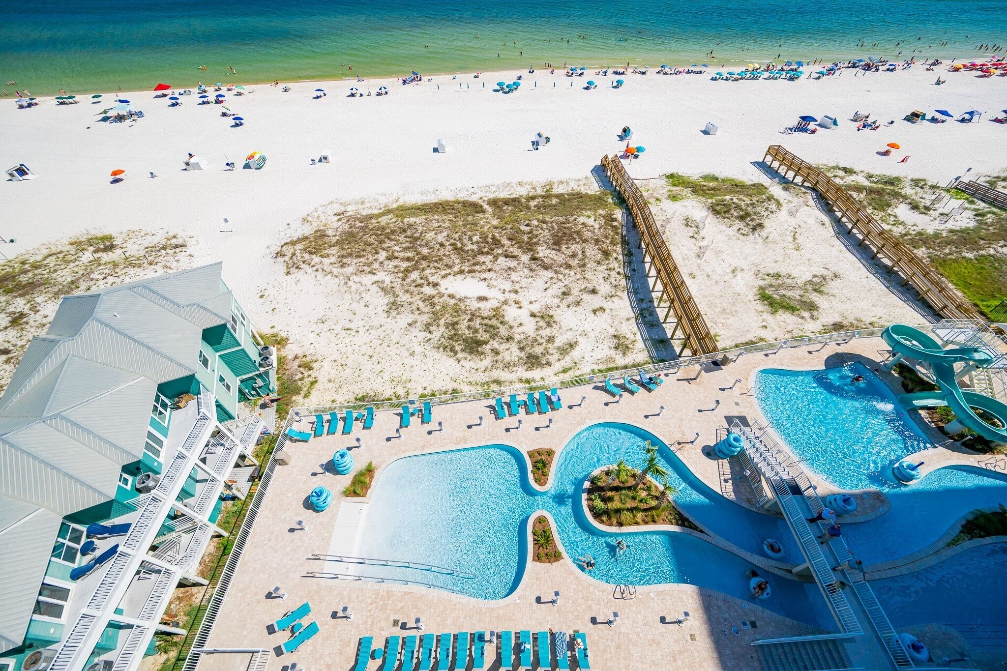An aerial view of a beach resort with a large pool area, a wooden boardwalk, and a beach with umbrellas and sun loungers.