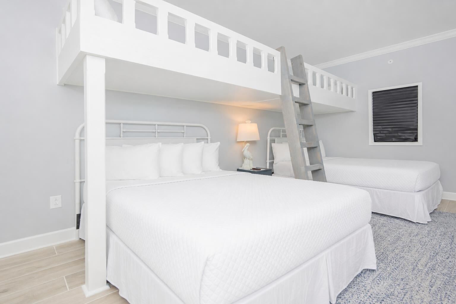 Une chambre avec un lit superposé, une échelle, une table de chevet avec une lampe et une fenêtre avec des stores.