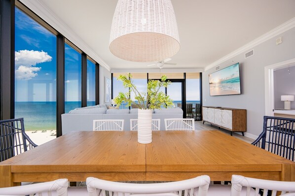 Un comedor moderno con una mesa de madera, sillas blancas, un florero blanco con flores y vista al océano a través de ventanas amplias.