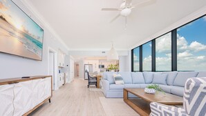 Una sala de estar moderna con un televisor grande, un sofá, una mesa de centro y vistas del cielo a través de amplias ventanas.