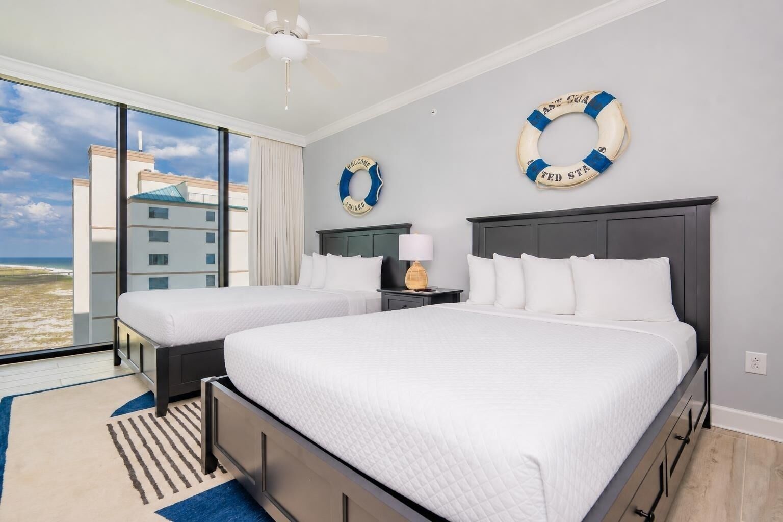 A hotel room with two beds, a view of the beach, a ceiling fan, and life rings on the wall.