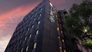 Ένα σύγχρονο κτίριο με μια μεγάλη, κάθετη επιγραφή που φέρει κείμενο, πολλά παράθυρα και ένα δέντρο στα δεξιά.