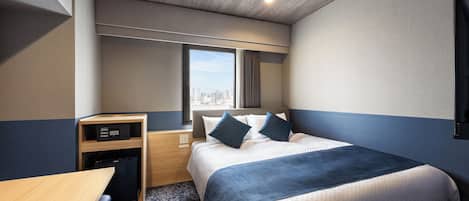 Habitación de hotel con cama, escritorio, refrigerador pequeño, ventana con vista a la ciudad e iluminación empotrada en el techo.