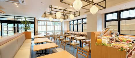Un comedor moderno con mesas y bancos de madera, ventanas amplias y una barra tipo buffet con comida y bebidas.
