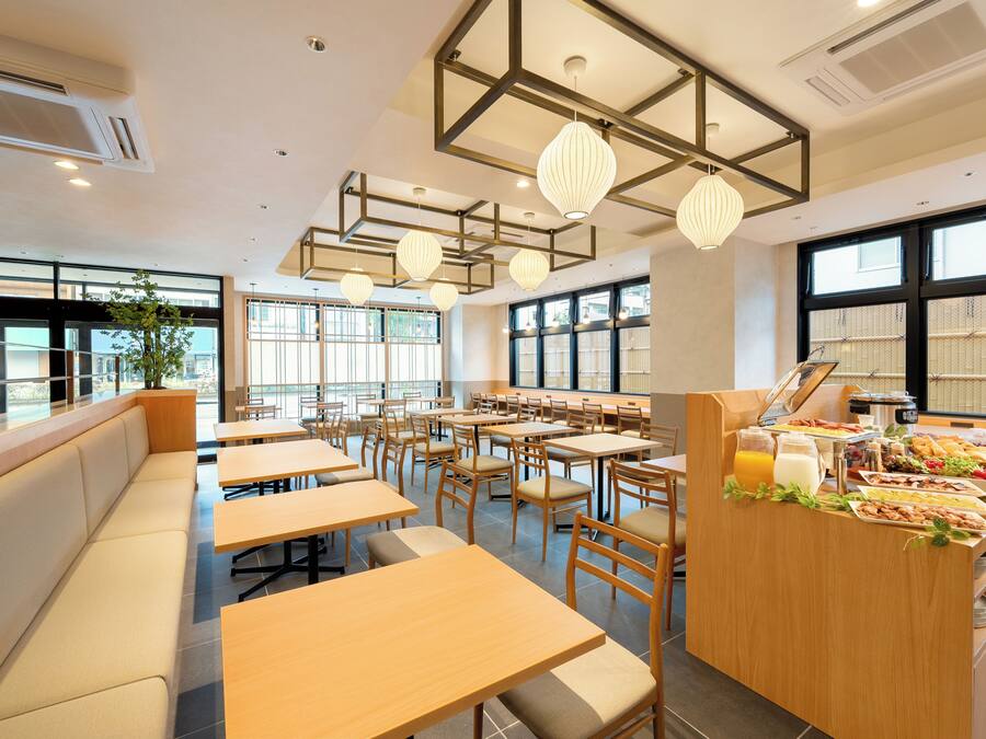 A modern dining area with wooden tables and benches, large windows, and a buffet counter with food and drinks.
