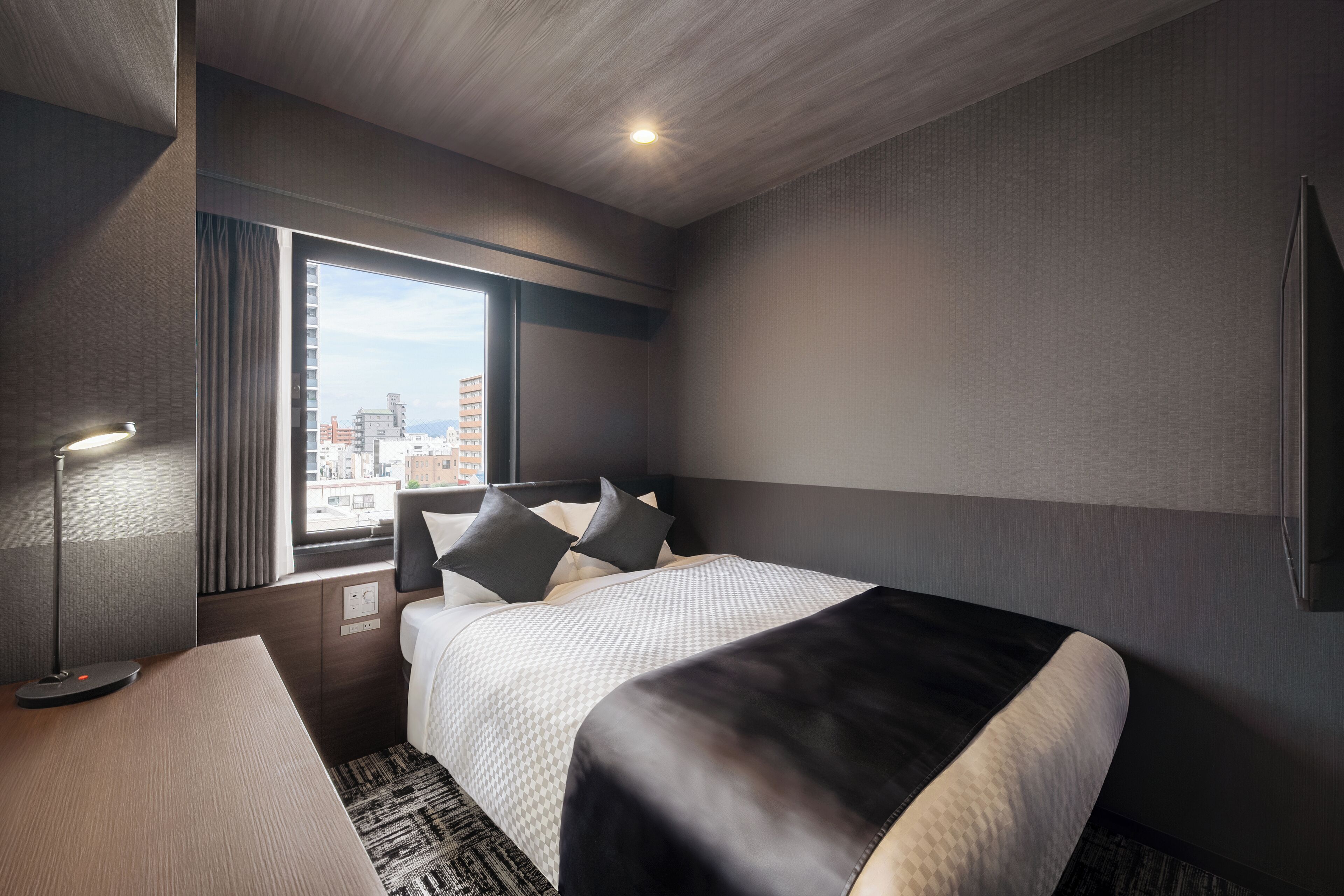 Une chambre d’hôtel moderne avec un grand lit, une table de chevet, une lampe et une fenêtre donnant sur la ville.