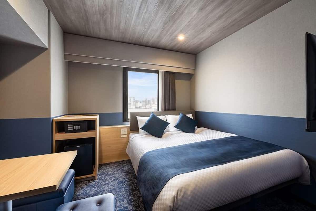 Habitación de hotel con cama, escritorio, refrigerador pequeño y ventana con vista a la ciudad.