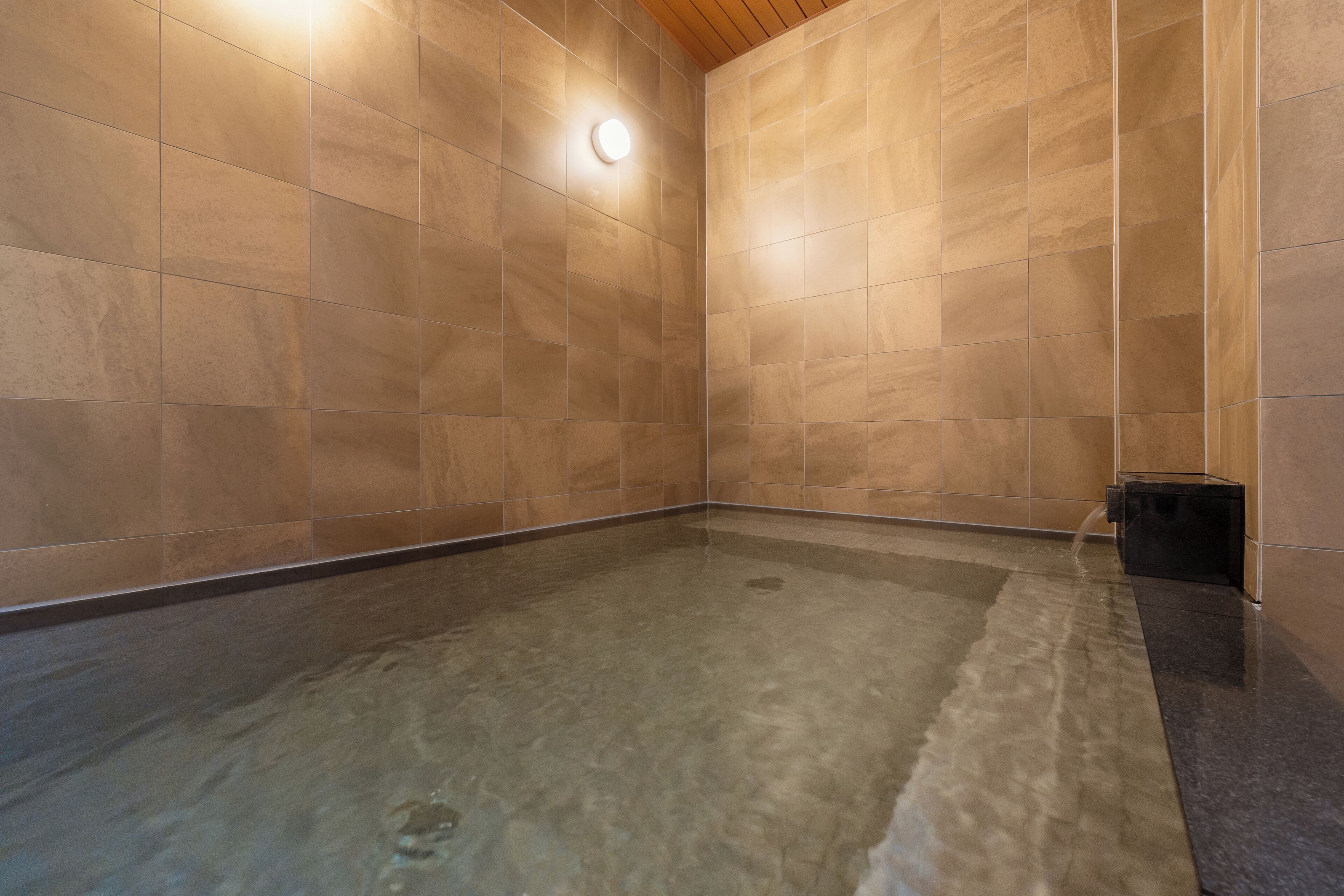 A spa room with a tiled floor and walls, a wooden ceiling, and a small black container on the right.