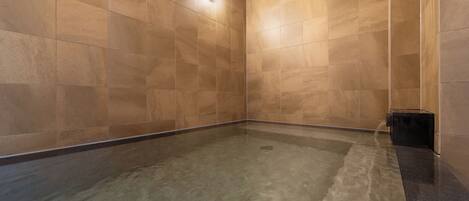 A spa room with a tiled floor and walls, a wooden ceiling, and a small black container on the right.