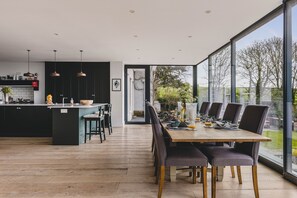 Een moderne keuken met een centraal eiland, een eetgedeelte met een grote tafel en stoelen, en grote ramen met uitzicht op de omgeving.