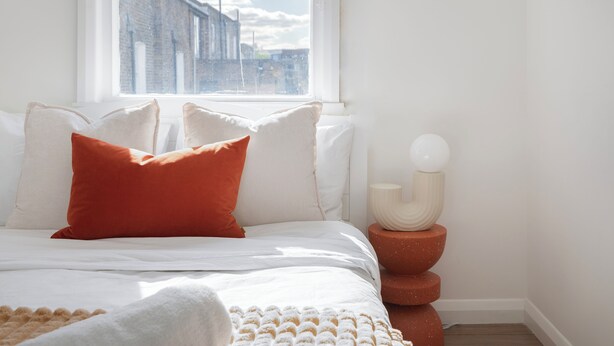 Ein ordentlich bezogenes Bett mit weißen und orangefarbenen Kissen, einer weißen Decke und einer gemusterten Tagesdecke. Auf einem Stapel Terrakottatöpfen neben dem Bett steht eine weiße Vase mit einem kugelförmigen Objekt.