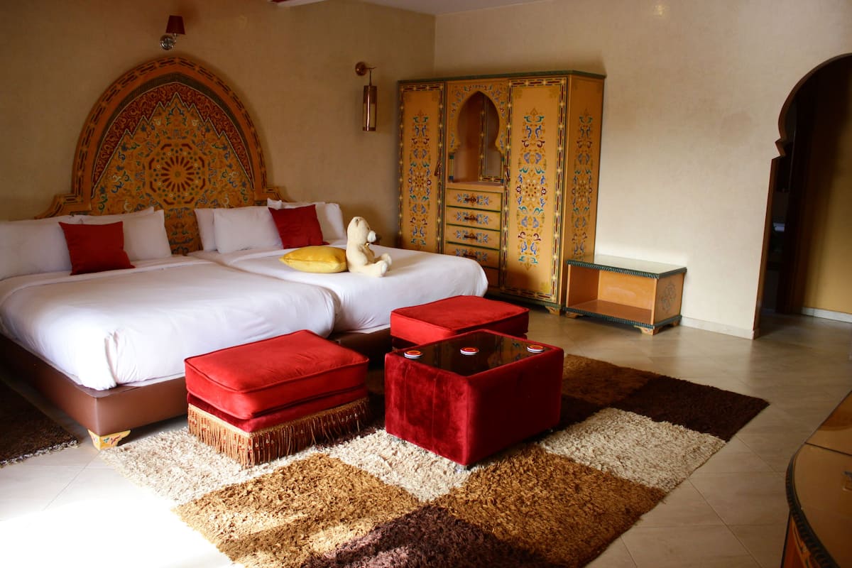 A bedroom with a large bed, two red ottomans, a decorative headboard, a wardrobe, and a small table.
