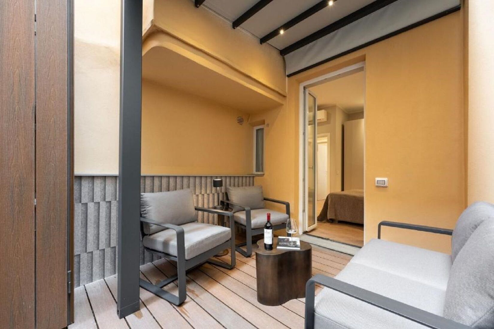 Una habitación moderna con un sofá, dos sillones, una mesita con una botella y un vaso, y una cama al fondo.