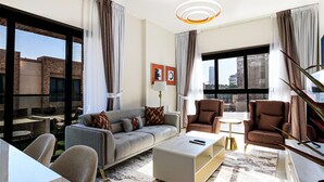 A modern living room with a sofa, armchairs, a coffee table, and a TV. There are large windows with curtains, a lamp, and artwork on the wall.