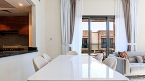 A modern living room with a dining table, sofas, and a kitchen area visible through a glass door.