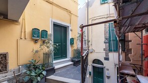 Une ruelle étroite avec un bâtiment jaune, des volets verts et une porte rouge. On y trouve des plantes en pot et une rambarde en métal.