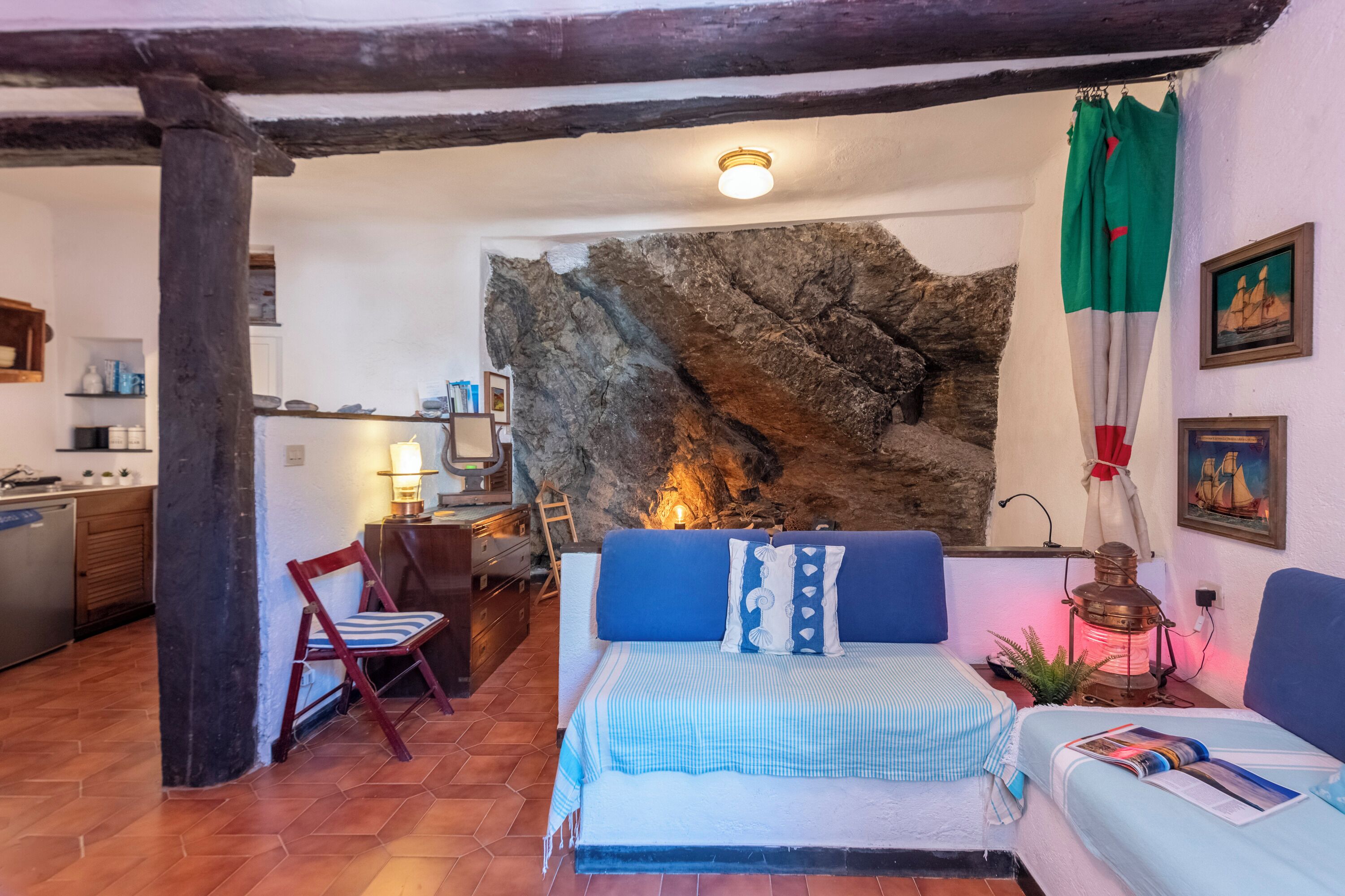 Habitació acollidora amb llit, cadira, taula i una cuina visible a través d'una porta oberta.