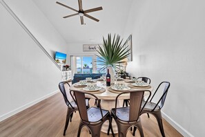 A dining area with a round table, chairs, a ceiling fan, and a view of the beach through the door.
