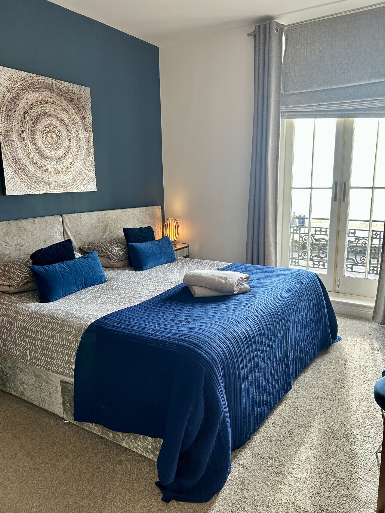 A bedroom with a large bed, a headboard, a bedside table with a lamp, and a view of the outside through a window.
