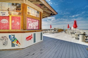 A wooden beach hut with a red sign reading 'WELCOME TO PELICAN BEACH TIKI HUT,' surrounded by beach chairs and umbrellas.