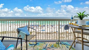 A balcony with a view of a beach, a table with a plant, and a chair with a cushion. - ☀️ Pelican Beach 603 - Starfish by the Sea - Gulf Views ☀️ (Destin)