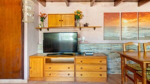 Un meuble TV en bois avec un téléviseur à écran plat, un buffet en bois avec des tiroirs, une table à manger avec des chaises, et un mur orné d’un tableau représentant un coucher de soleil.