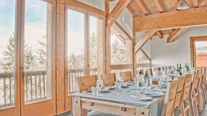Un comedor con una larga mesa de madera dispuesta para una comida, ventanas grandes y vistas a árboles.