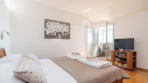 Ein ordentlich gemachtes Bett mit einem Kissen mit Punkten, einer beigen Decke und gefalteten Handtüchern. An der Wand hängt ein Blumenbild, ein Fernseher steht auf einem Holzgestell und eine Glastür führt zu einem Balkon.