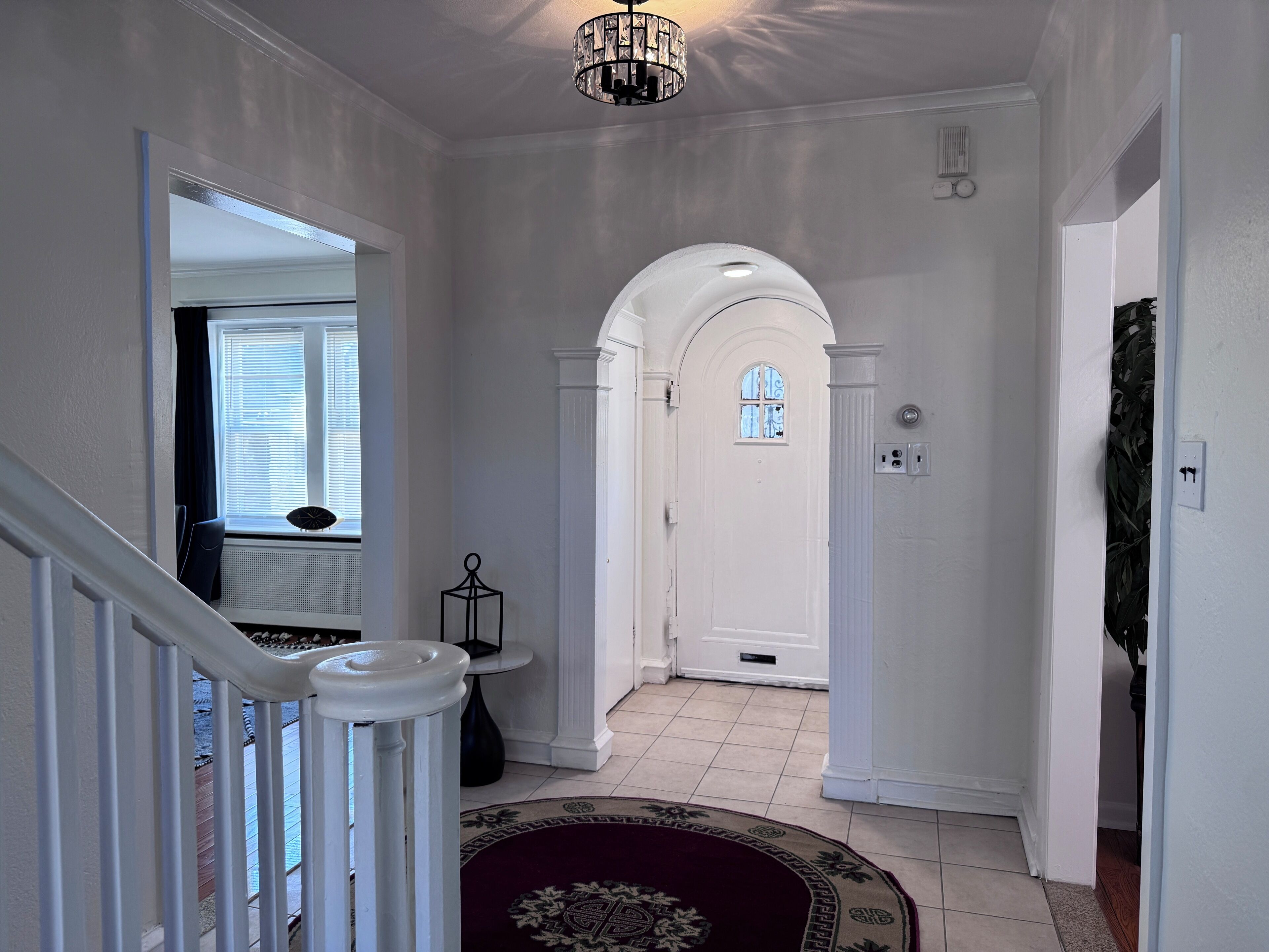Un couloir aux murs blancs, une porte blanche avec une fenêtre, un lustre et une petite table avec une lampe.