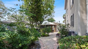 A garden pathway leading to a wooden gazebo, surrounded by lush greenery and a house with a gabled roof.