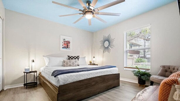 Ein Schlafzimmer mit einem Bett, einem Deckenventilator, einem Fenster mit Jalousien, einem Sessel und einer Pflanze.