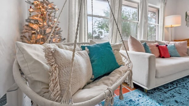 Un salon chaleureux avec un canapé blanc, un fauteuil suspendu agrémenté de coussins, un sapin de Noël et une grande fenêtre avec des rideaux.