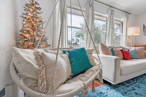 A cozy living room with a white sofa, a hanging chair with cushions, a Christmas tree, and a large window with curtains. - Pink House in Prime Location Dogs Ok Walk to DT (St. Augustine)