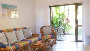 Une pièce comprenant un canapé, des fauteuils, une petite table et donnant sur un espace extérieur aménagé avec des plantes, visible à travers une porte vitrée.