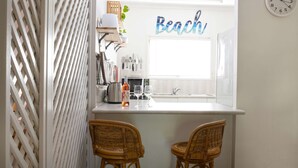 Une cuisine avec un plan de travail blanc, deux chaises en osier, une bouteille de vin et un panneau décoratif représentant une plage accroché au mur.