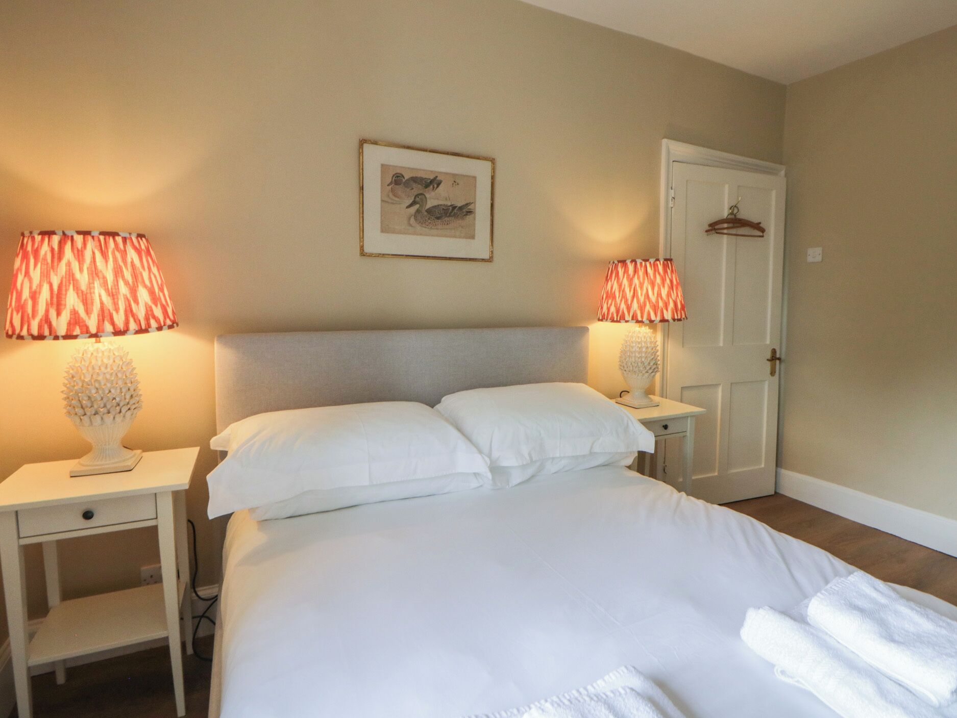 A bedroom with a bed, two bedside tables, a lamp, a door, and a wall-mounted picture.