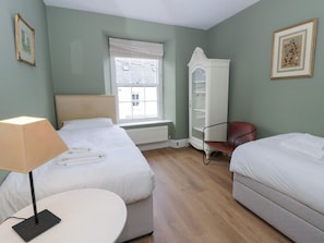 A room with two beds, a chair, a table, a radiator, a window, and a cabinet.