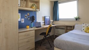 Et soveværelse med seng, skrivebord med computer, stol og et vindue med gardiner.
