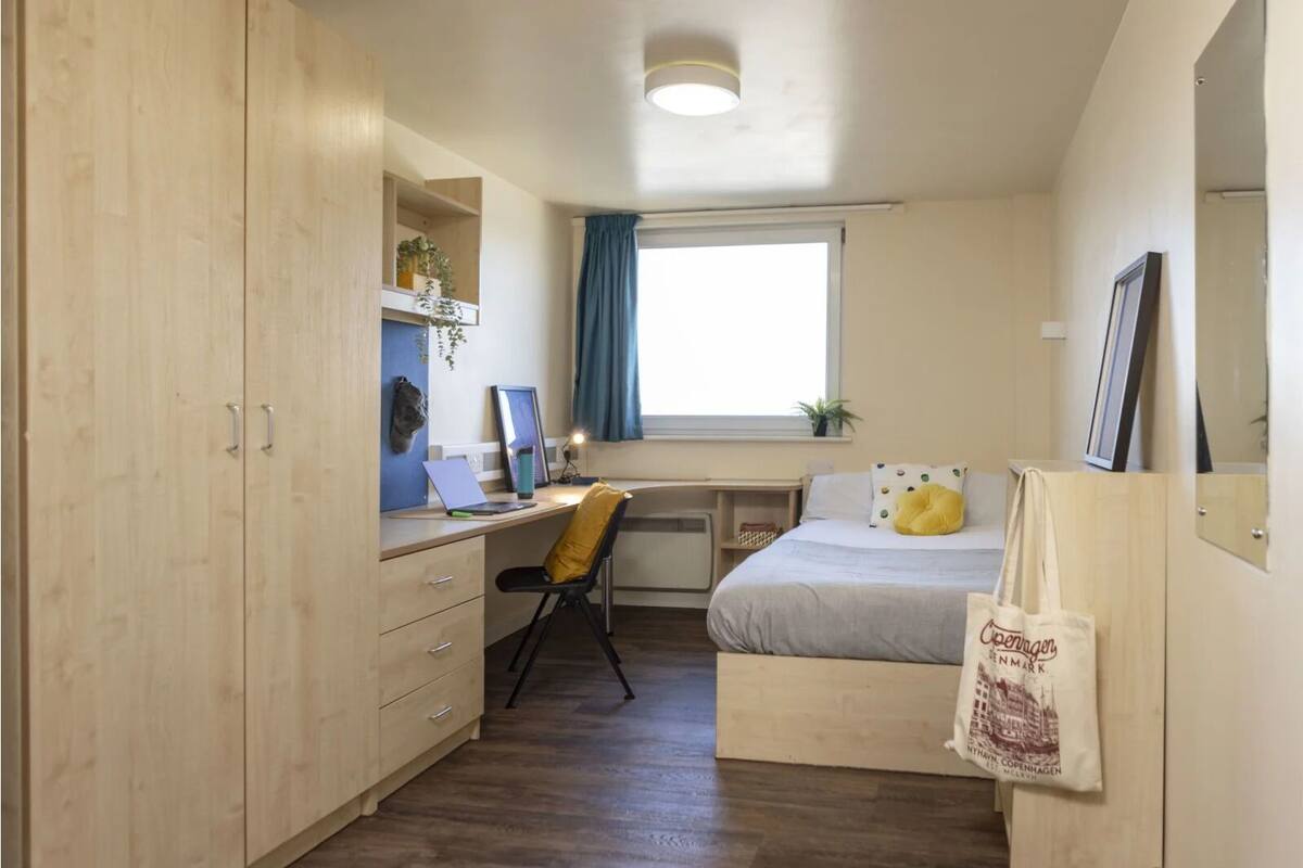 A compact bedroom with a wooden wardrobe, a desk with a chair, a bed with a pillow, and a window with curtains.