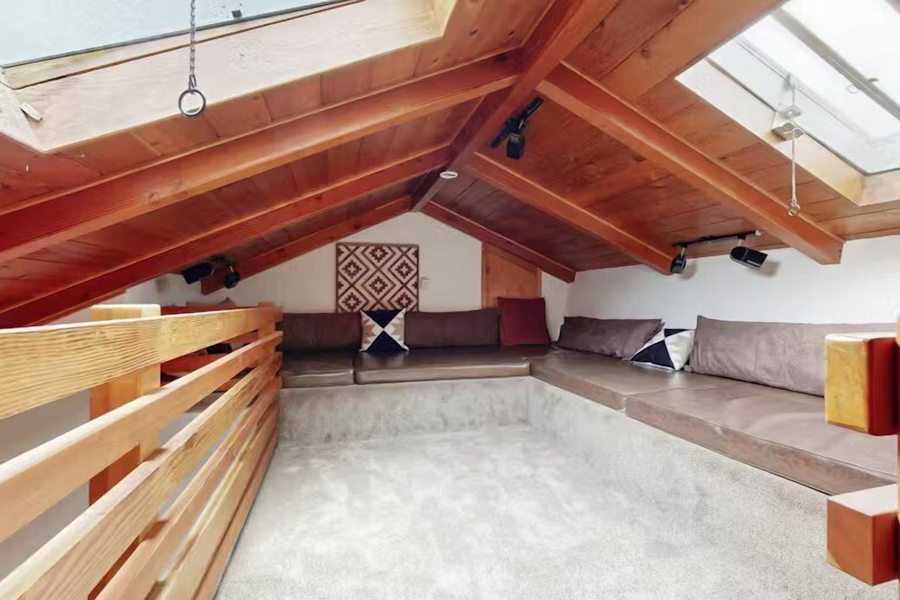 Techo inclinado con vigas de madera, un área de descanso con cojines y un decorado de pared con estampado.