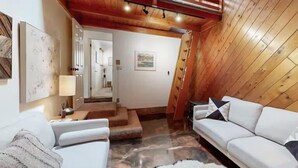 A cozy, well-lit room with a wooden ceiling, white sofas, a staircase, and a view of a mountain.
