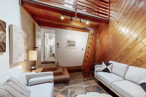 A cozy, well-lit room with a wooden ceiling, white sofas, a staircase, and a view of a mountain.