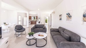 A spacious living area with a grey sofa, a round coffee table, and a grey armchair. There is a dining area with white chairs and a table, and a kitchen in the background.