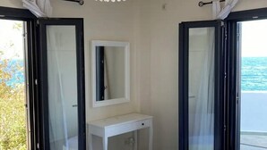 Een slaapkamer met een bed, een witte nachtkastje, een spiegel en een plafondlamp. Twee schuiframen leiden naar een balkon met uitzicht op zee.