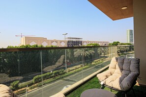 A balcony with a glass barrier, a cushioned chair, and a view of a road with palm trees and construction cranes in the background.