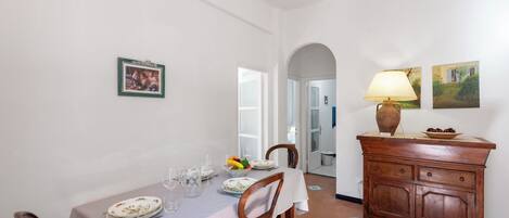 Un coin repas avec une table dressée pour deux personnes, des chaises, un meuble avec une lampe et un tableau encadré au mur.
