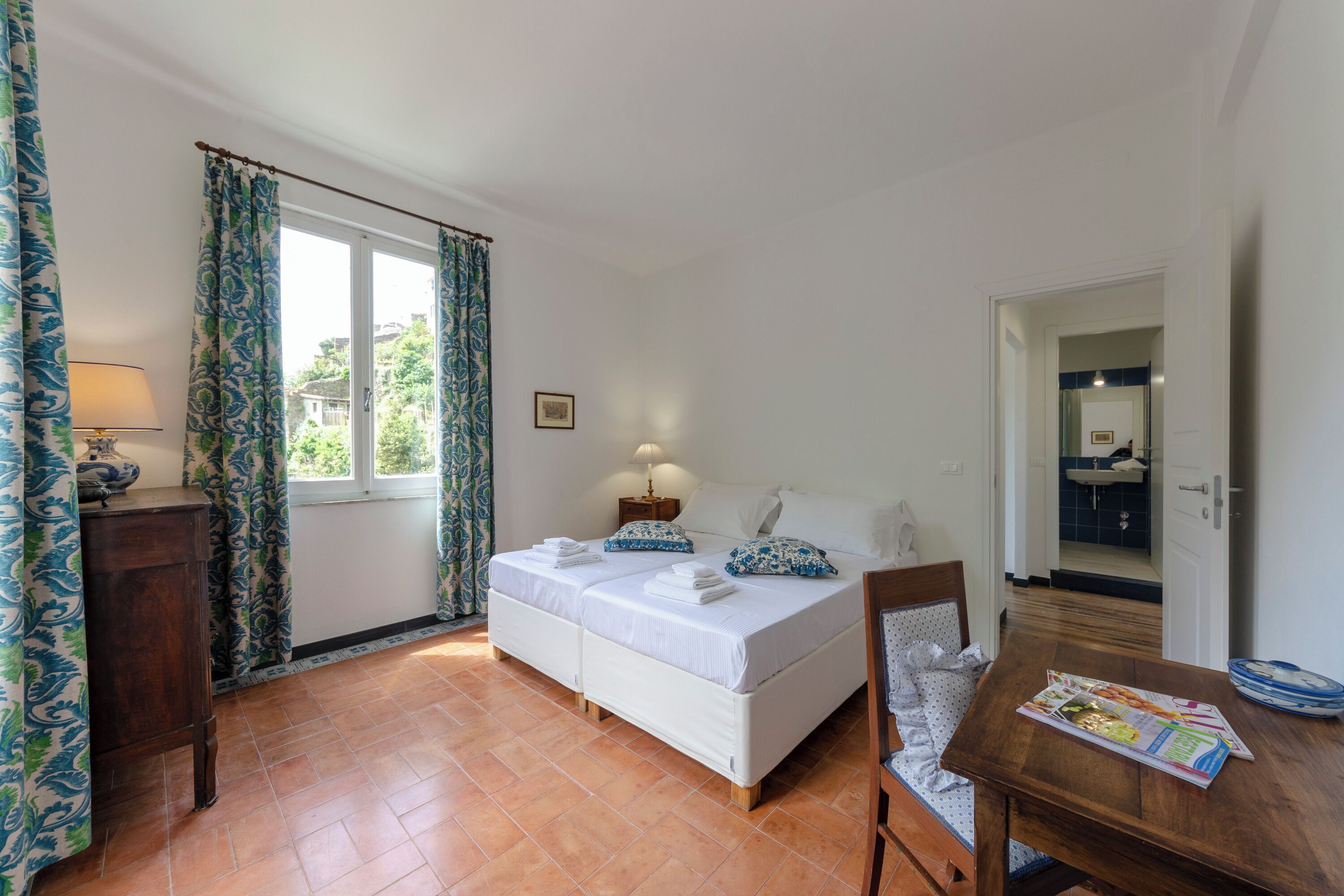 Une chambre à coucher avec un lit double, une table de chevet en bois, une petite table de chevet avec une lampe, et une vue sur l’extérieur par une fenêtre avec des rideaux.