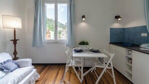 Habitación pequeña con mesa de comedor, sillas blancas, lámpara, ventana con cortinas y vistas a edificios.