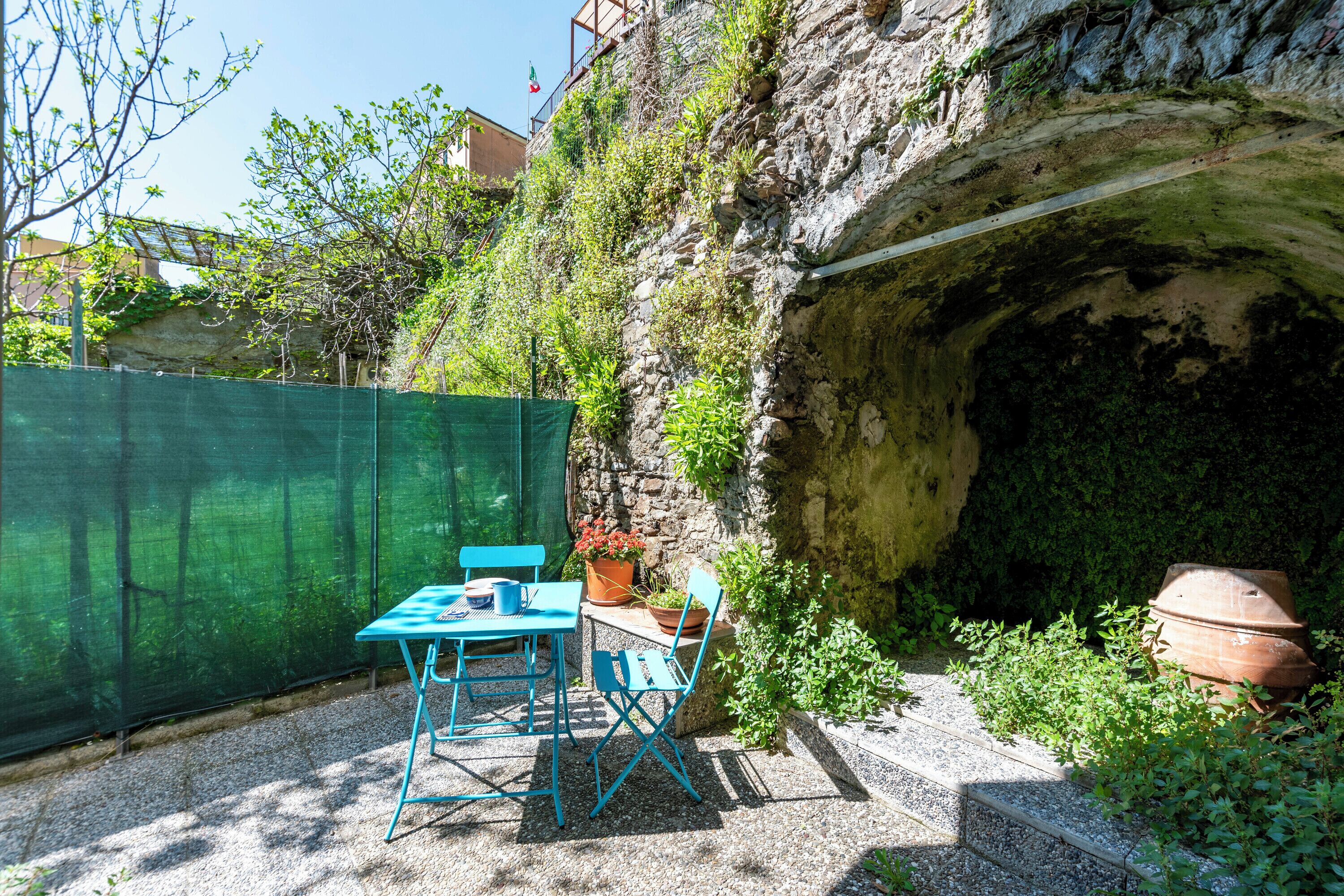 A patio with a table and chairs, a green fence, and a rocky wall with a tunnel entrance.