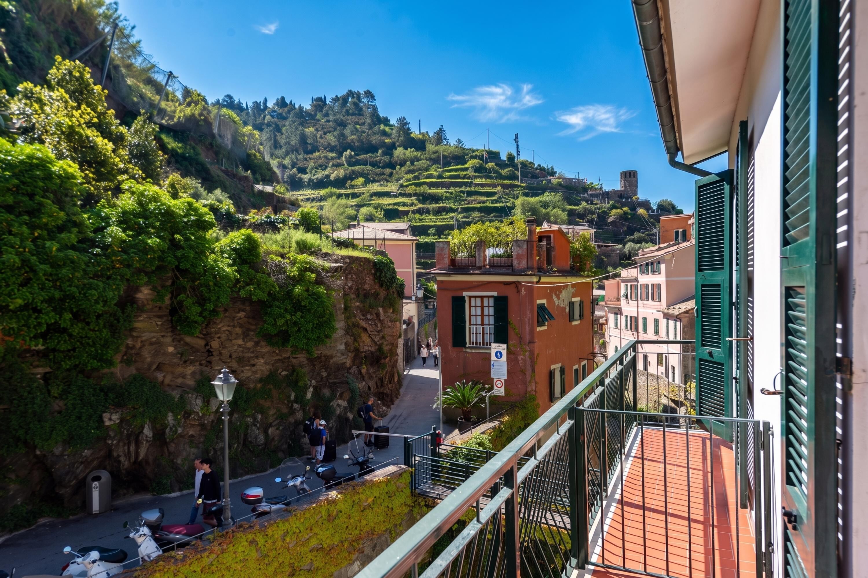 A balcony with a view of a hillside, a street with parked scooters, and buildings with traditional architecture.